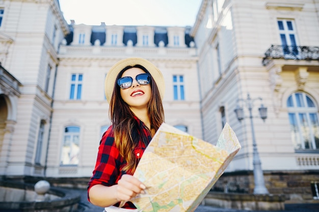 Chica turista feliz con cabello castaño con sombrero y camisa roja, sosteniendo el mapa en el fondo de la antigua ciudad europea y sonriendo, viajando, retrato.