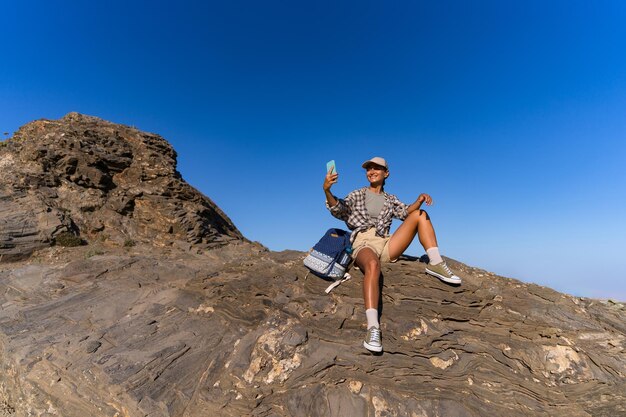 una chica turista con una mochila en el verano en la cima de una montaña toma una selfie contra el telón de fondo