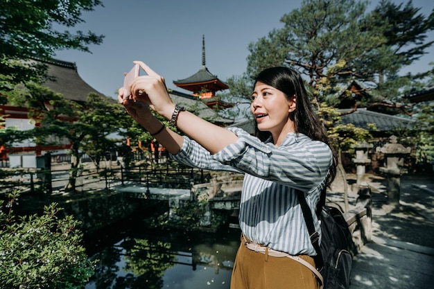 chica turista sosteniendo una aplicación de cámara de teléfono celular tomando fotos de hermosos paisajes en el jardín japonés cerca del santuario. una joven mochilera asiática usando un teléfono inteligente toma una foto bajo el sol kiyomizu dera