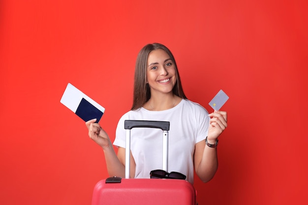 Chica turista en verano mostrando tarjeta de crédito plástica, con gafas de sol, maleta roja, pasaporte aislado sobre fondo rojo.