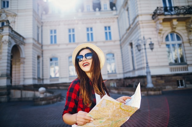 Chica turística atractiva con cabello castaño con sombrero, gafas de sol y camisa roja, sosteniendo el mapa en el fondo de la antigua ciudad europea y sonriendo, viajando, retrato.