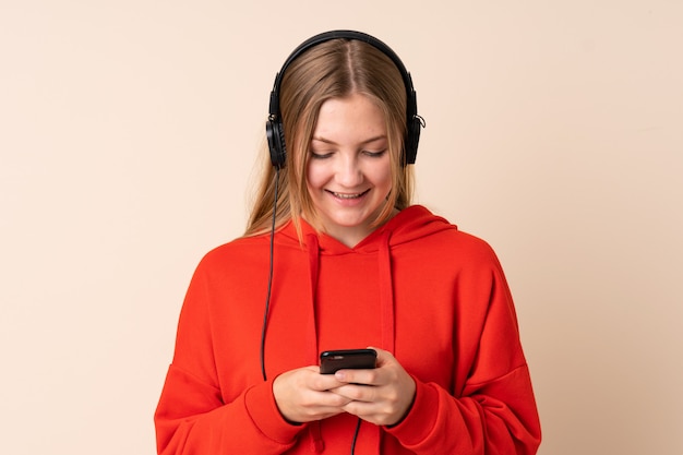 Chica ucraniana adolescente aislada en beige escuchando música y mirando al móvil