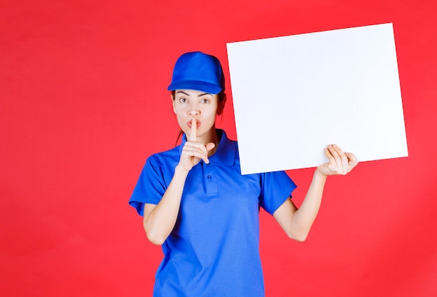 Foto chica en uniforme azul y boina sosteniendo un mostrador de información cuadrado blanco y pidiendo silencio.