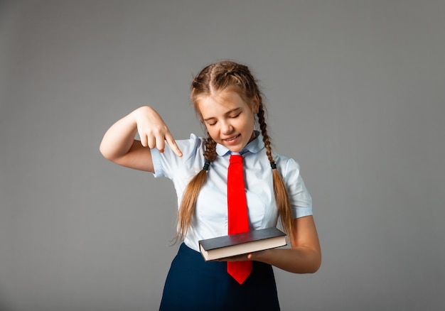 Chica en uniforme escolar sosteniendo un libro en sus manos con emociones pensativas en su rostro, posando sobre un fondo gris