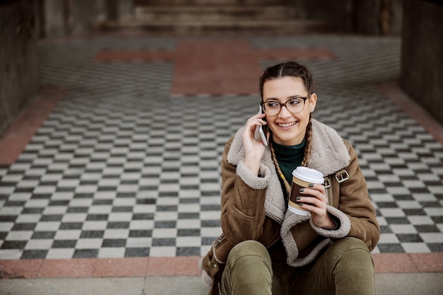Chica universitaria alegre sentada en la entrada del edificio de la universidad, conversando por teléfono y sosteniendo una taza desechable con café.