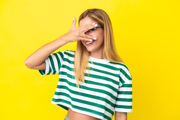 Chica uruguaya rubia aislada de fondo amarillo cubriendo los ojos con las manos y sonriendo