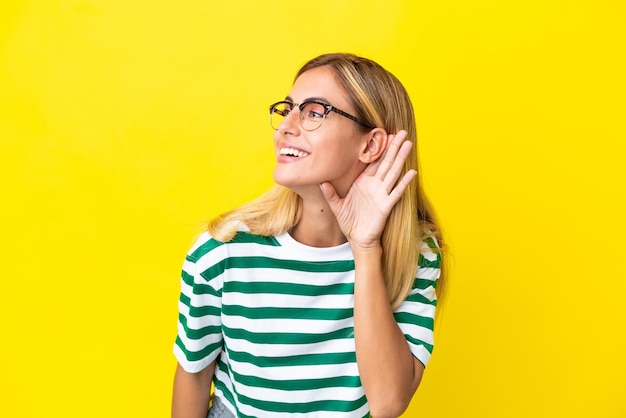 Chica uruguaya rubia aislada de fondo amarillo escuchando algo poniendo la mano en la oreja