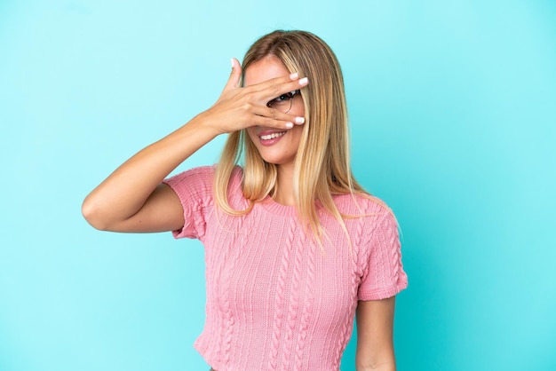 Chica uruguaya rubia aislada de fondo azul cubriendo los ojos con las manos y sonriendo