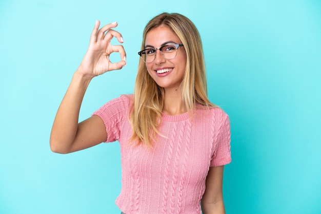 Chica uruguaya rubia aislada de fondo azul que muestra el signo de ok con los dedos