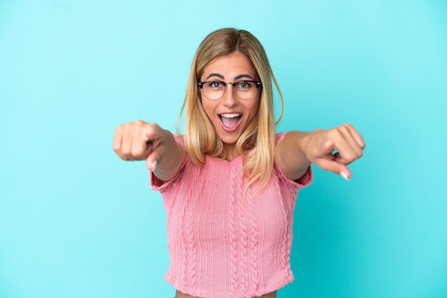 Chica uruguaya rubia aislada de fondo azul sorprendida y apuntando al frente