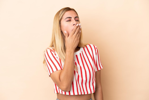 Chica uruguaya rubia aislada de fondo beige bostezando y cubriendo la boca abierta con la mano