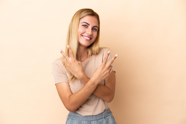 Chica uruguaya rubia aislada de fondo beige sonriendo y mostrando el signo de la victoria