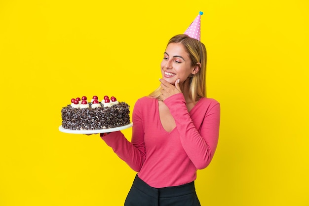 Chica uruguaya rubia sosteniendo pastel de cumpleaños aislado de fondo amarillo mirando hacia un lado y sonriendo