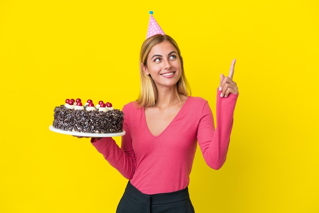 Chica uruguaya rubia sosteniendo pastel de cumpleaños aislado de fondo amarillo señalando una gran idea