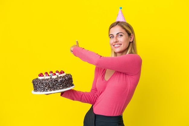 Chica uruguaya rubia sosteniendo pastel de cumpleaños aislado sobre fondo amarillo apuntando hacia atrás