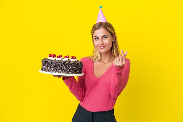 Chica uruguaya rubia sosteniendo pastel de cumpleaños aislado sobre fondo amarillo haciendo gesto de dinero