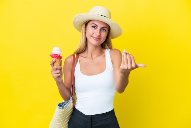 Chica uruguaya rubia en verano sosteniendo helado aislado en un fondo amarillo invitando a venir con la mano Feliz de que hayas venido