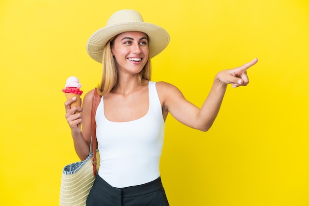 Chica uruguaya rubia en verano sosteniendo helado aislado en fondo amarillo señalando con el dedo al costado y presentando un producto