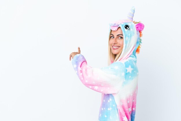 Foto chica uruguaya rubia vistiendo un pijama de unicornio aislado sobre fondo blanco apuntando hacia atrás