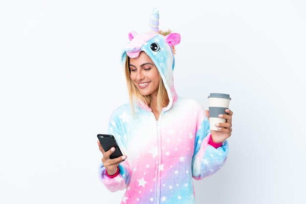 Chica uruguaya rubia vistiendo un pijama de unicornio aislado sobre fondo blanco sosteniendo un café para llevar y un móvil