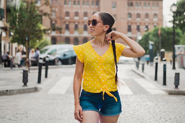 Chica de vacaciones caminando por las calles de Madrid día soleado vistiendo una camiseta amarilla y pantalones cortos de mezclilla gafas de sol cabello en una larga trenza