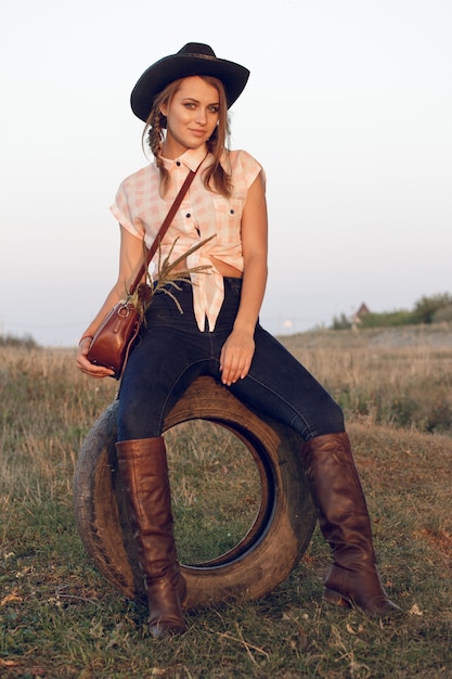 Foto chica vaquera en camisa de botas de jeans y con una bolsa en una caja con una rueda por un árbol