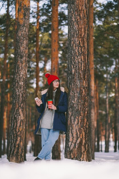 Chica con vaso de papel y un teléfono en un bosque nevado