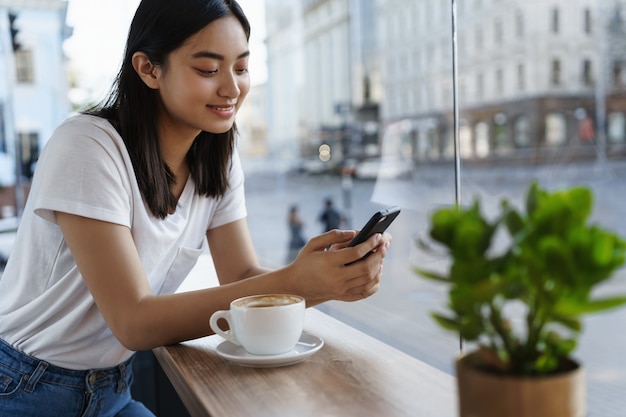 Chica de verano sentada en la cafetería con una taza de café, mensajería en el teléfono inteligente.