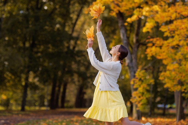 Chica vestida de amarillo en el parque de otoño se regocija en otoño sosteniendo hojas amarillas en sus manos cálidas