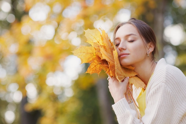Chica vestida de amarillo en el parque de otoño se regocija en otoño sosteniendo hojas amarillas en sus manos cálidas