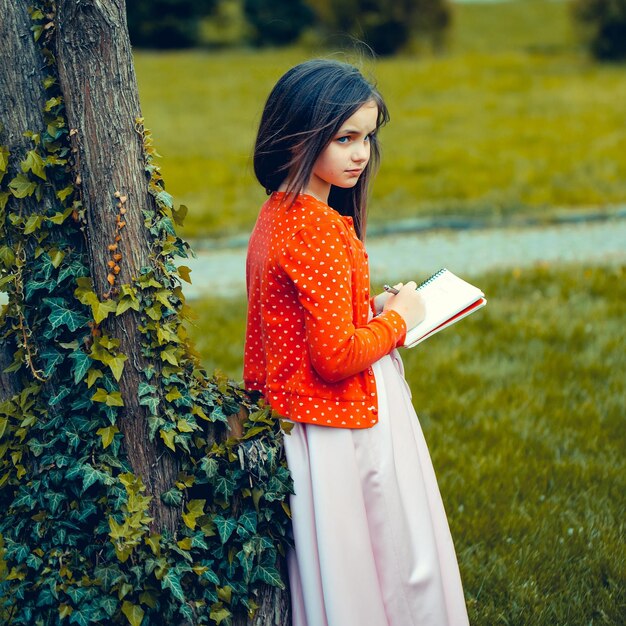 Chica vestida con cuaderno y bolígrafo cerca del árbol.