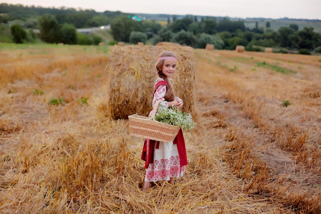 una chica vestida de eslavo con una cesta de margaritas en las manos se encuentra en un campo junto a un pajar