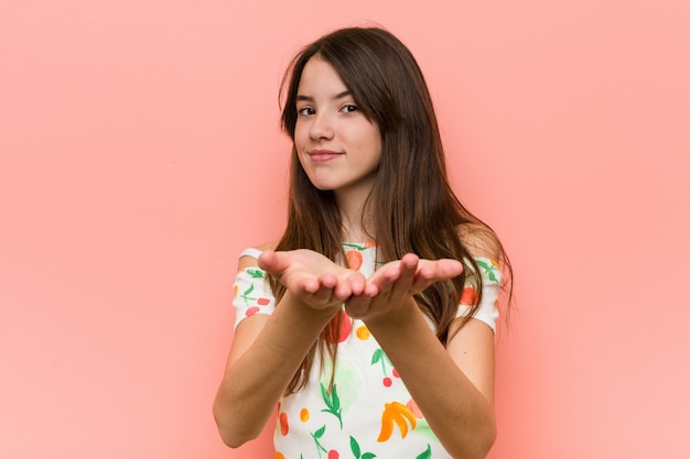 Chica vestida con una ropa de verano contra una pared rosa sosteniendo algo con palmeras, ofreciendo a la cámara