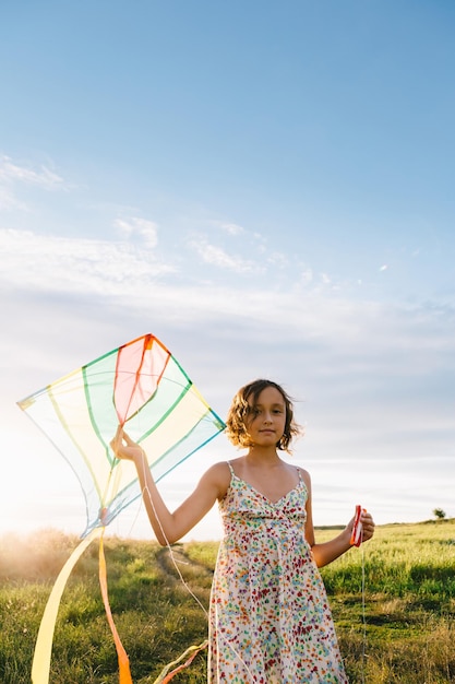 Chica vestida sosteniendo una cometa brillante mientras corre y mira hacia otro lado en un campo verde de verano