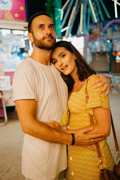 Una chica con un vestido amarillo con un escote pronunciado y su novio con barba se abrazan entre atracciones. Una pareja de enamorados en una cita en la feria de Valencia.