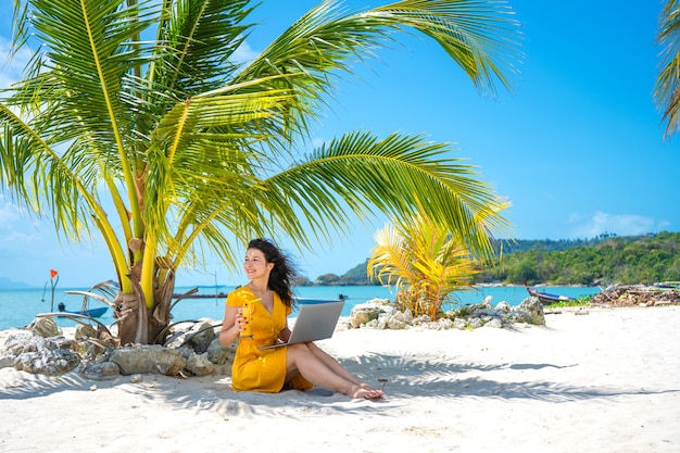 Foto chica con un vestido amarillo en una playa tropical de arena trabaja en una computadora portátil y bebe mango fresco. trabajo remoto, freelance exitoso. trabaja en vacaciones.