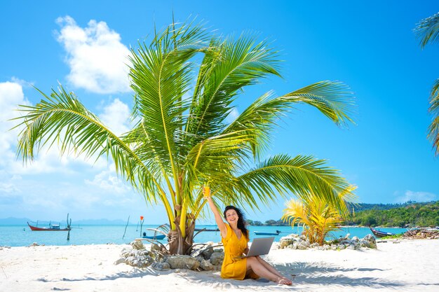 Chica con un vestido amarillo en una playa tropical de arena trabaja en una computadora portátil y bebe mango fresco. Trabajo remoto, freelance exitoso. Trabaja en vacaciones.