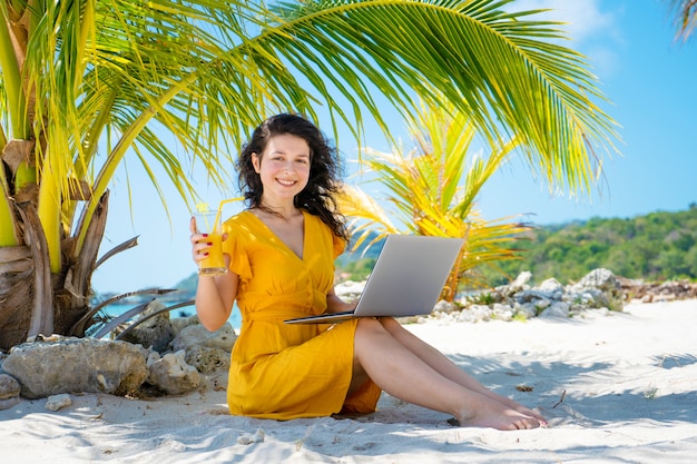 Chica con un vestido amarillo en una playa tropical de arena trabaja en una computadora portátil y bebe mango fresco. Trabajo remoto, freelance exitoso. Trabaja en vacaciones.