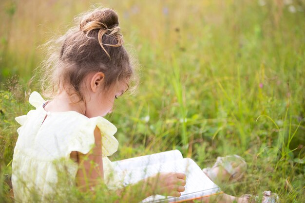 Chica con un vestido amarillo se sienta en la hierba sobre una manta en un campo y lee un libro de papel. Día Internacional del Niño. Horario de verano, infancia, educación y entretenimiento, núcleo rural. Copia espacio