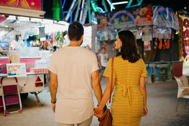 Una chica con un vestido amarillo y su novio de espaldas caminan entre atracciones. Una pareja de enamorados en una cita en la feria de Valencia.