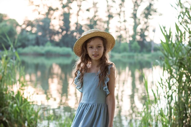 Una chica con vestido azul y sombrero de paja se encuentra en la orilla del lago.