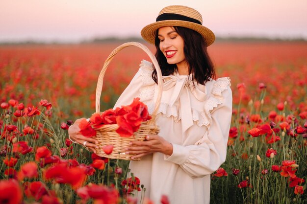 Una chica con un vestido blanco y una cesta de amapolas camina por un campo de amapolas