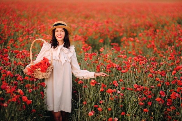 Una chica con un vestido blanco y una cesta de amapolas camina por un campo de amapolas