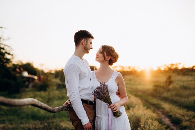 Chica con un vestido blanco y un chico con una camisa blanca en un paseo al atardecer con un ramo de flores en un pueblo fuera de la ciudad