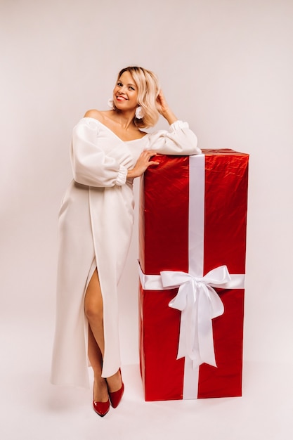 Una chica con un vestido blanco con un enorme regalo rojo sobre un fondo blanco sonríe.