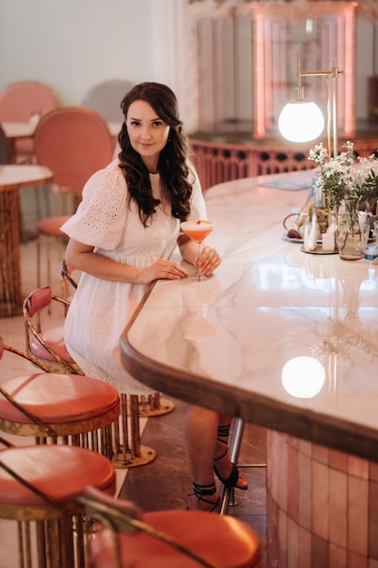 Una chica con un vestido blanco está sentada en el bar de un café y bebiendo un cóctel.