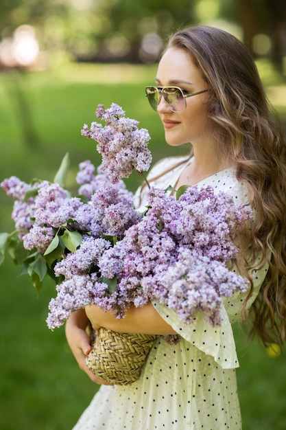 La chica de vestido blanco y gafas de sol sostiene en sus manos una canasta de mimbre con flores. Cesta con lilas. Niña y flores. Camina con una canasta de lilas en las manos. Florística