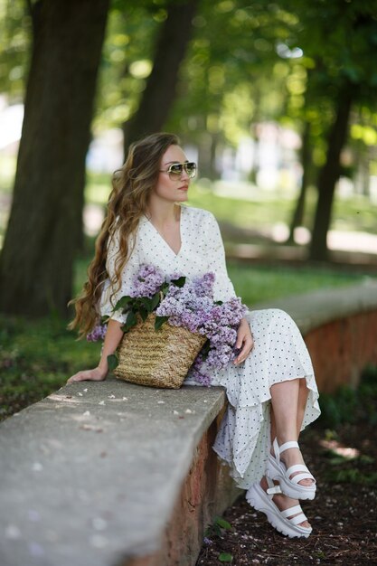 La chica de vestido blanco y gafas de sol sostiene en sus manos una canasta de mimbre con flores. Cesta con lilas. Niña y flores. Siéntese con una canasta de lilas en las manos. Florística