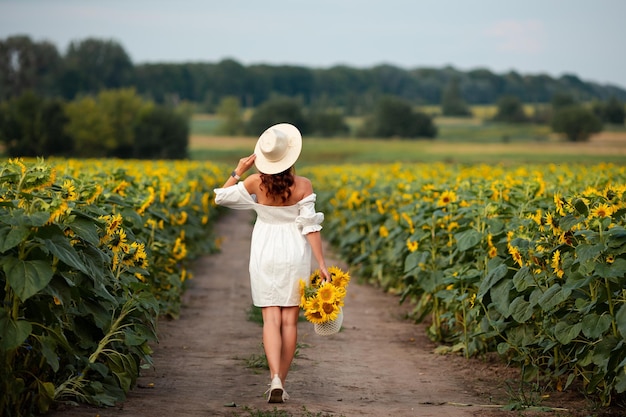 Una chica con un vestido blanco y un sombrero camina por un campo de girasoles.