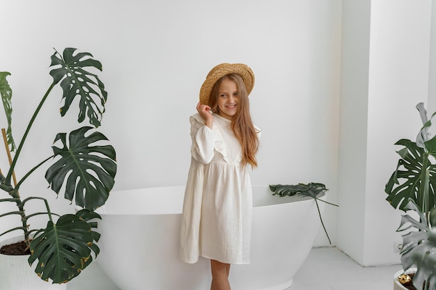 una chica con un vestido blanco y un sombrero de paja en una habitación con plantas y baño